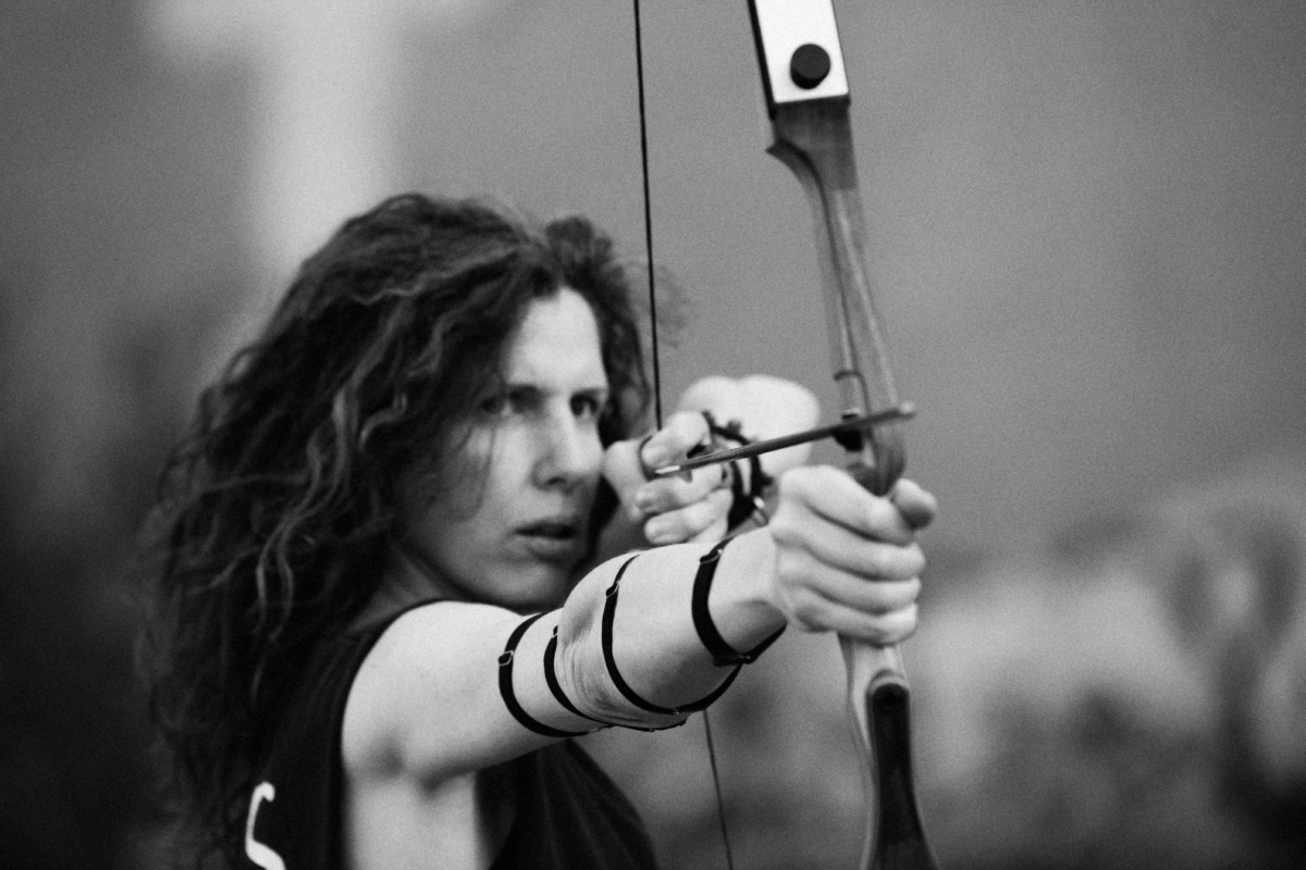 A black and white photo of Aisha with a bow and arrow drawn. She's has long dark curly hair and is calmly staring down the shaft of the arrow like she's about to fuck something up.